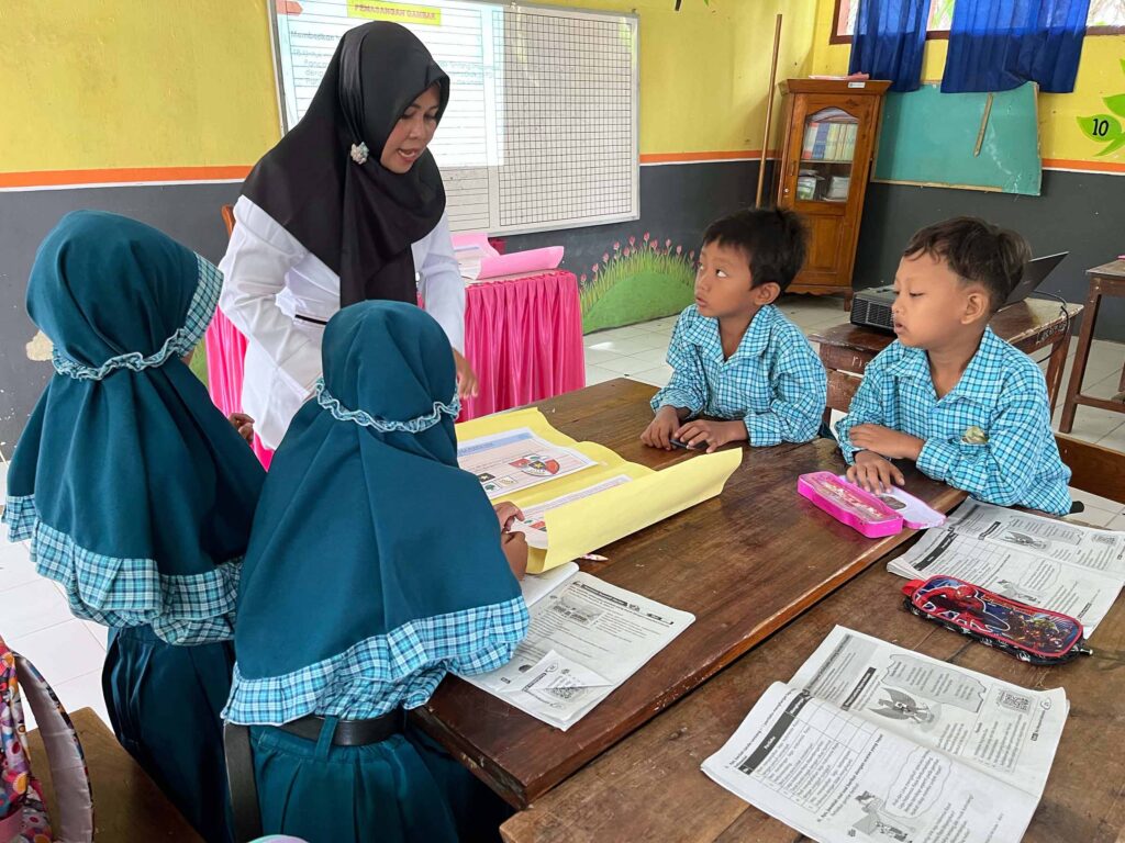 A female teacher wearing a black headscarf stands over a small group of four elementary school students—two boys and two girls in blue headscarves—seated at a wooden table in a classroom. The students are working on a lesson, illustrating the impact of STIR Education teaching methodologies implemented through the STIR Program Indonesia.