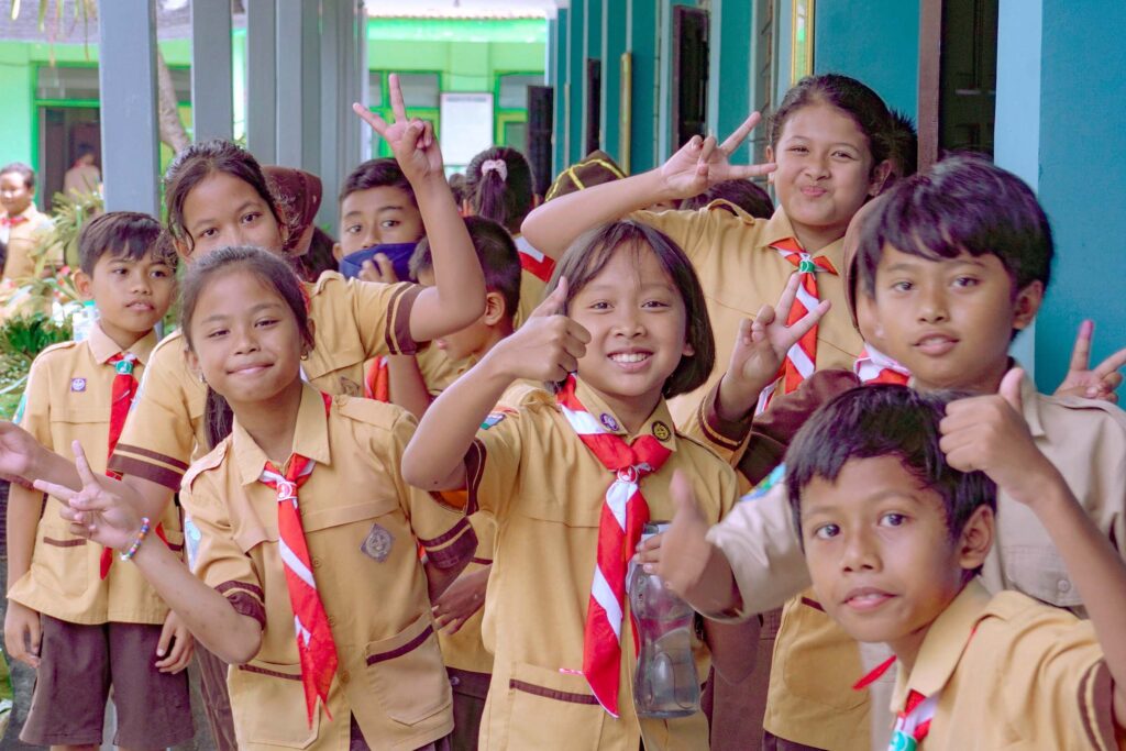 A large group of cheerful Indonesian elementary school students, wearing their light brown and orange Scout (Pramuka) uniforms with red and white scarves, are posing for a group photo outdoors at school. Many of the children are smiling and enthusiastically making peace signs and thumbs-up gestures, representing the positive energy and engagement fostered by STIR Education within the STIR Program Indonesia.