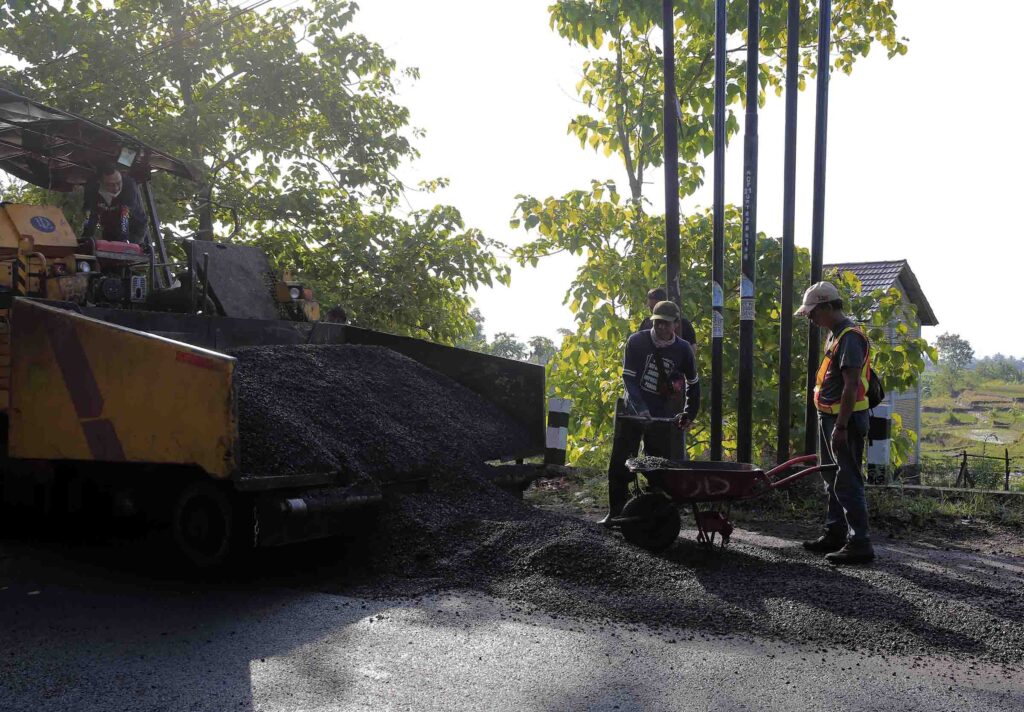 Construction workers standing next to an asphalt paver machine, using a wheelbarrow to move the hot asphalt mix for placement on the road.