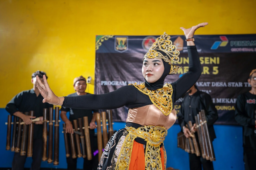 An elderly man wearing a traditional headband guides a teenage boy's hand as he strikes a large hanging gong with a mallet.