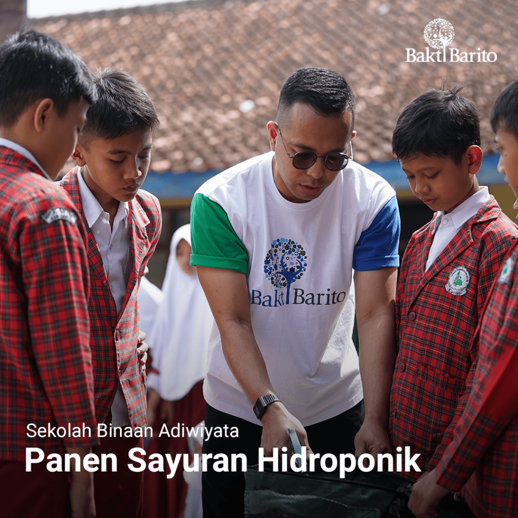 A group photograph outdoors where a male mentor, Rinaldi Dwinata Simatupang wearing a Bakti Barito t-shirt with a blue sleeve and green sleeve, points at a seedling tray for four young students. Below the image, bold white text reads: 'Yayasan Bakti Barito & Chandra Asri Berdayakan Masyarakat Lewat Pelatihan Pertanian Hidroponik.