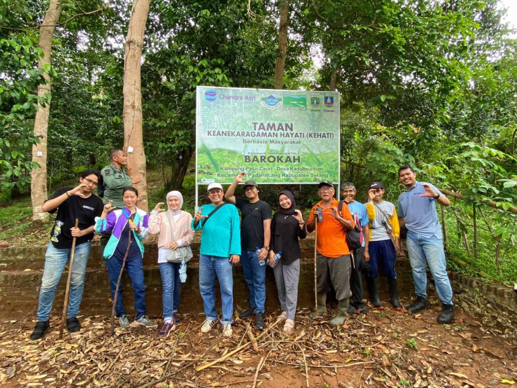 Chandra Asri Group Taman Kehati Biodiversity Park Program: Eleven people of Chandra Asri employees, forest rangers, park security, and local community members standing in front of a large sign, Taman Keanekaragaman Hayati (KEHATI) for the Barokah Forest Farmers Group in Padarincang, Serang. The group is smiling and posing, with most making a "C" hand gesture.