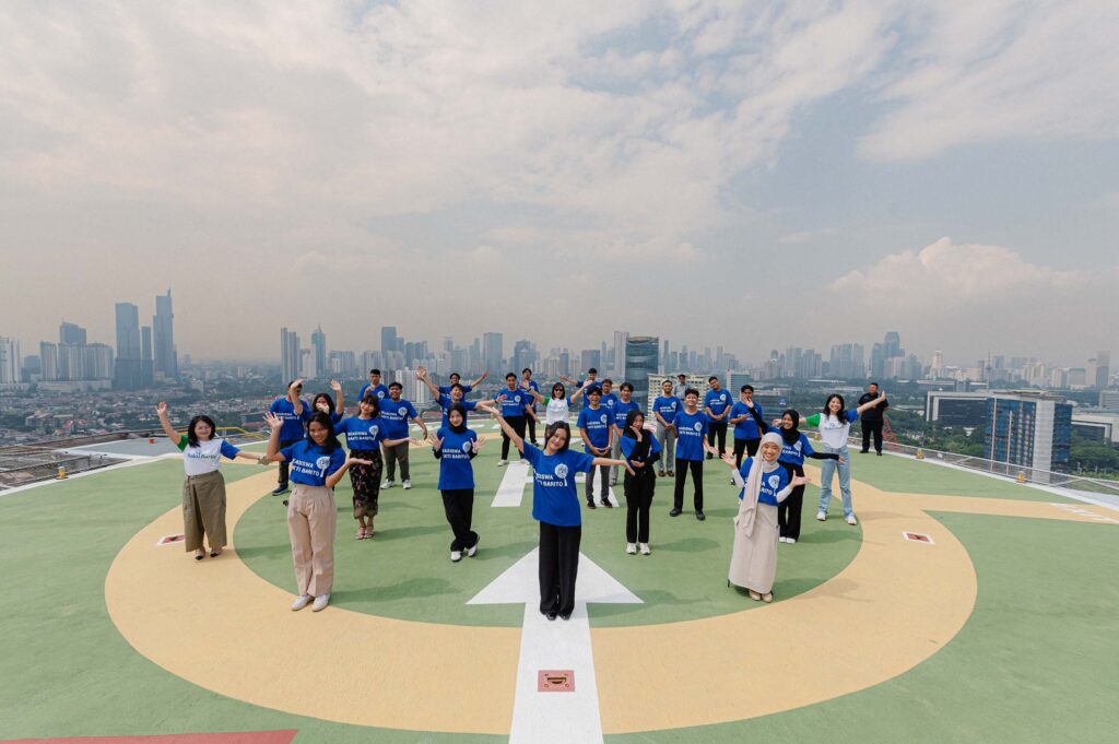 Bakti Barito Scholarship Program: A group of young adults in blue and white t-shirts stand in a formation on a green and yellow helipad with a sprawling city skyline in the background.