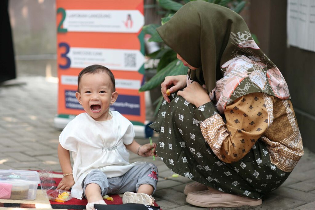 Bakti Barito Partnership to Accelerate Stunting Reduction in Indonesia (PASTI) Program: A young child sits smiling on an outdoor mat while a woman in an olive green hijab crouches beside them, both participating in a community nutritional support event